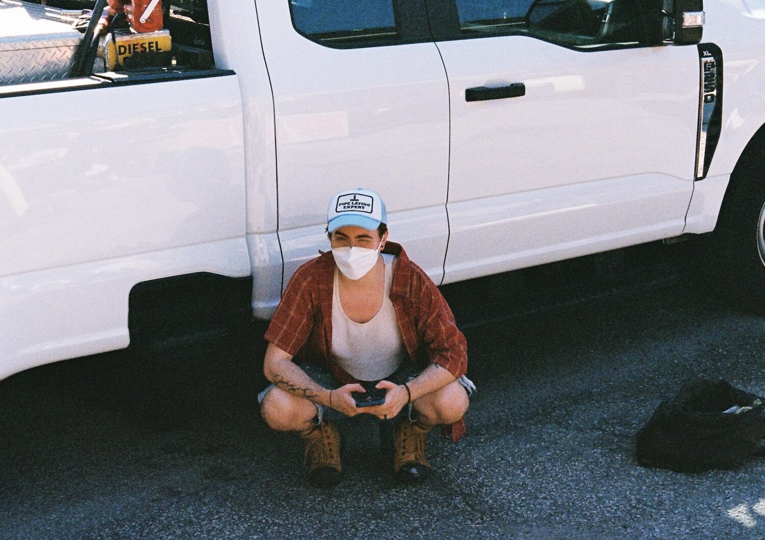 Antel squatting beside white pick-up truck on the trades-themed shoot, wearing a white KN95 mask and a blue trucker hat that says "pipe-laying expert."